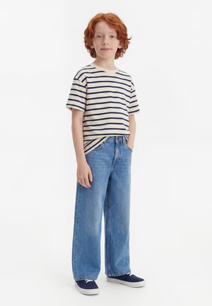 Enfant roux debout, portant un t-shirt à manches courtes rayé bleu marine et blanc, un jean bleu ample et des baskets bleu marine aux semelles blanches.
