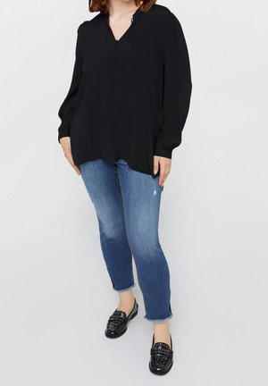 Kobieta w luźnej czarnej bluzce, niebieskich skróconych dżinsach z postrzępionymi brzegami i czarnych mokasynach z ćwiekami, stojąca na jednolitym jasnym tle.