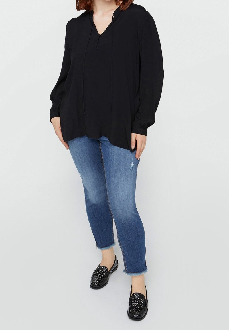Femme portant un chemisier noir ample, un jean bleu court avec des ourlets effilochés et des mocassins noirs cloutés, debout devant un fond clair uni.