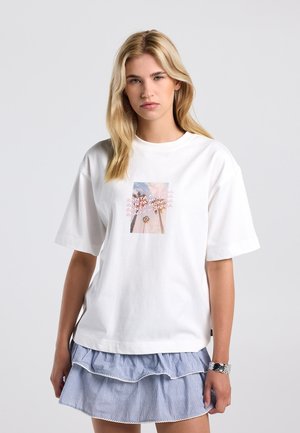 Blonde vrouw draagt een wit T-shirt met een Santa Monica-graphic en een blauwe gestreepte gelaagde rok, staand tegen een effen achtergrond.