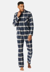 Pijamas de franela a cuadros en azul marino y blanco, que incluyen una camisa con botones y un bolsillo en el pecho, junto con pantalones a juego. Tejido suave y texturizado.