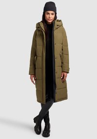 Long olive green puffer coat with detachable hood, zip closure, side pockets, and quilted texture; worn over a black outfit and boots.