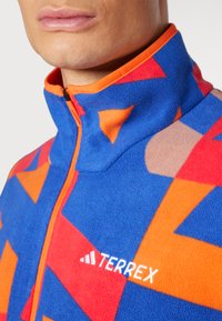 Fleecejakke i blå med orange geometriske mønstre, høj krave og lynlåslukning. Har hvidt logo på brystet.