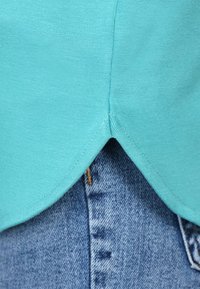 Türkises Baumwollhemd mit gewelltem Saum, kombiniert mit hellblauer Jeans, die sichtbare Nähte und einen Knopfverschluss aufweist.