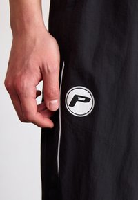 Čierne atletické nohavice majú biely kruhový logotyp s písmenom "P". Materiál vyzerá ľahko a má viditeľnú štruktúru. Prítomné sú aj biele akcentové lemovanie.