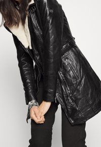 Chaqueta de cuero negra con cuello de borreguillo, que presenta varios bolsillos con cremallera y una cintura ajustada. Textura suave y diseño entallado.