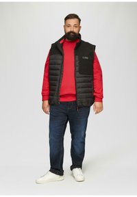 Gilet en duvet noir avec une fermeture éclair sur le devant et des poches latérales, porté sur un sweat-shirt rouge. Un jean en denim bleu et des baskets blanches complètent la tenue.