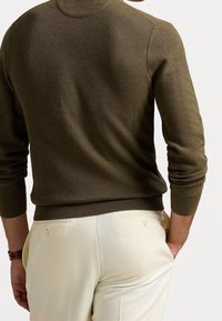 Olivgrön stickad tröja med hög krage, ribbade ärmslut och fåll, bärs över ljusa byxor med knäppta fickor.