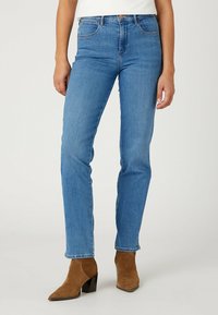Vaqueros de mezclilla azul con un ajuste slim, cintura de altura media, diseño de cinco bolsillos y un ligero desvanecimiento. Combinados con botas de ante marrón hasta el tobillo.