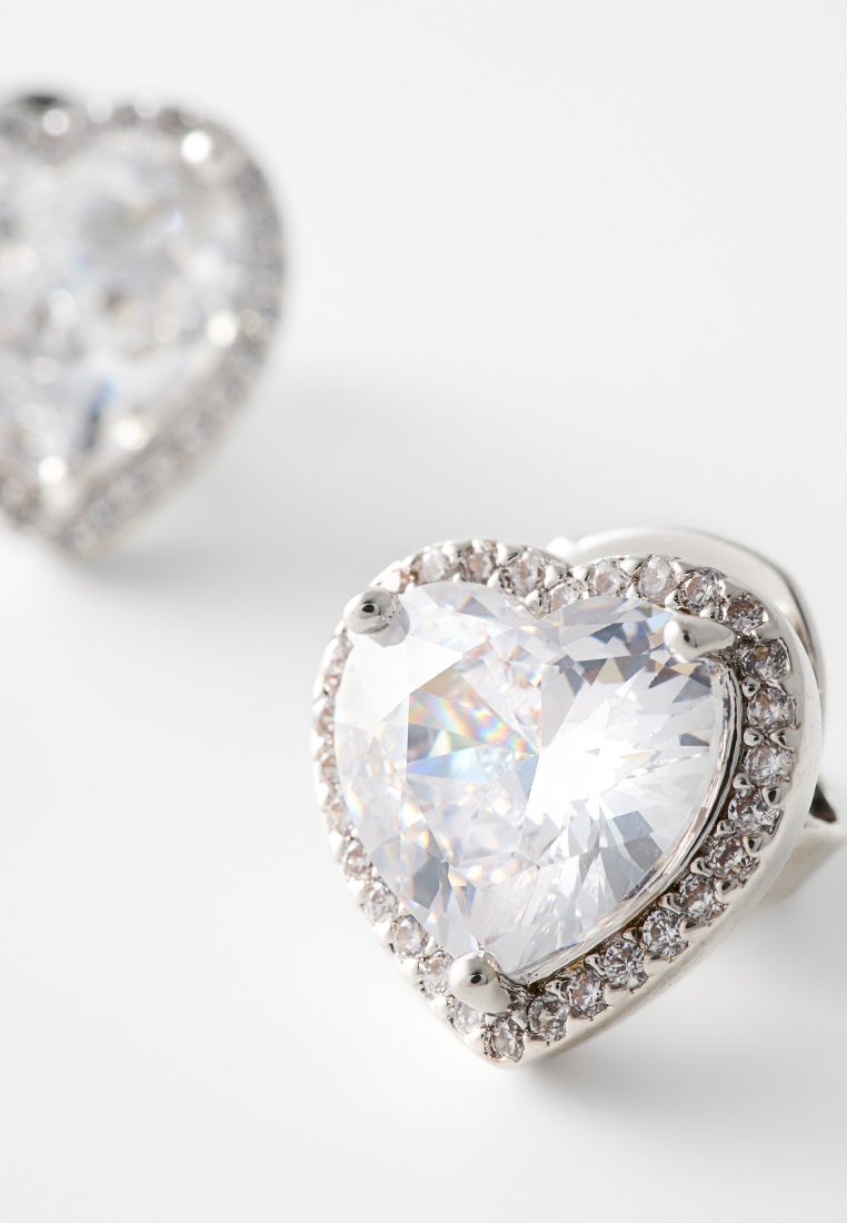 Sercowate kolczyki z dużym przezroczystym kryształem w centrum, otoczonym mniejszymi błyszczącymi kamieniami, osadzonymi w metalowej oprawie w srebrnym odcieniu.