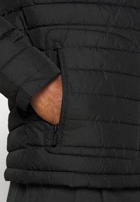 Chaqueta negra acolchada con un bolsillo lateral con cremallera. El material es texturizado, con un acabado ligeramente brillante y costuras horizontales.