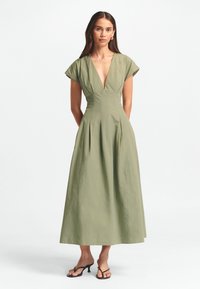 Oliivivihreä polvimittainen mekko, jossa on syvä V-pääntie ja tyllihihat. Mekossa on pleatitus vyötäröllä ja sileä, teksturoidun pinnan kangas.