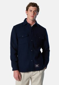Veste chemise en laine bleu marine avec deux poches poitrine, fermeture à boutons et un patch de marque en bas. Texture lisse et design structuré.