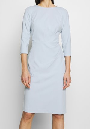 Femme portant une robe bleu clair, longueur genou, manches trois-quarts et taille cintrée, debout devant un fond blanc.
