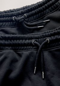 Gros plan d'une ceinture élastique noire sur un pantalon de survêtement avec des cordons noirs terminés par des œillets métalliques argentés.