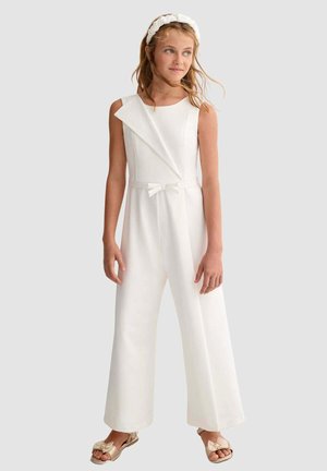 Ragazza che indossa una tuta bianca senza maniche con gambe larghe, un piccolo fiocco in vita, sandali di raso e una fascia bianca arricciata per capelli.