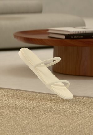 Μπεζ μινιμαλιστικό σανδάλι με δύο λεπτά λουράκια, τοποθετημένο πάνω σε υφασμένο χαλί, με στρογγυλό ξύλινο τραπεζάκι και στοίβες βιβλίων στο παρασκήνιο.