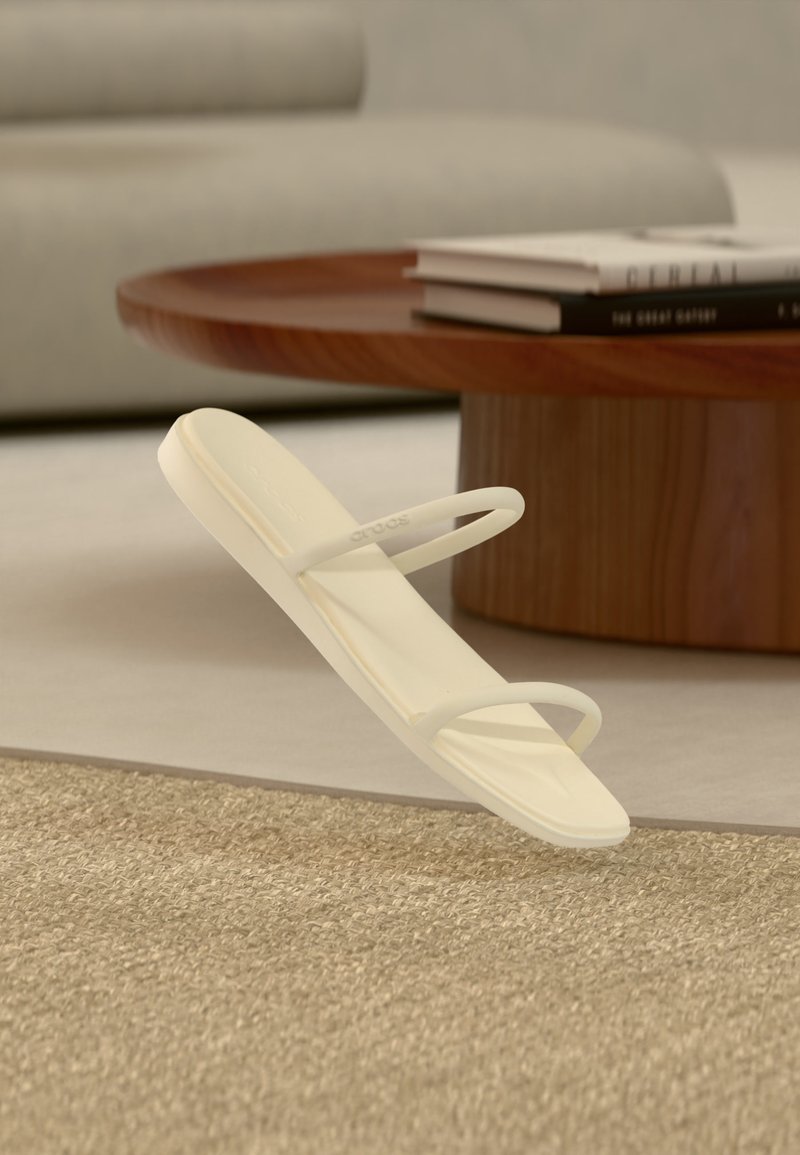 Μπεζ μινιμαλιστικό σανδάλι με δύο λεπτά λουράκια, τοποθετημένο πάνω σε υφασμένο χαλί, με στρογγυλό ξύλινο τραπεζάκι και στοίβες βιβλίων στο παρασκήνιο.