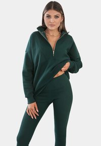 Groene rits-sweater met ribbeltextuur en lange mouwen, gecombineerd met bijpassende ribbel leggings. Heeft een hoge kraag en zijsplitten.