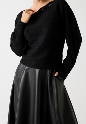 Kvinde iført en sort strikket sweater med ballonarme og et sort lædernederdel, den ene hånd rører kraven og den anden i lommen.