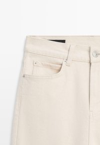 Primo piano di jeans in denim color crema che mostrano la tasca anteriore, l'asola per la cintura, i rivetti in metallo e la chiusura con bottone.