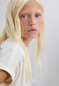 Top beige claro con detalles fruncidos en los hombros. La modelo tiene el cabello rubio lacio y largo y ojos azules, con un fondo neutro.