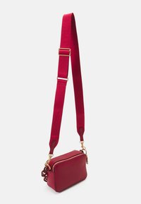 Sac bandoulière en cuir rouge avec fermeture à zip, doté d'une large sangle ajustable et de quincaillerie dorée. Texture lisse avec des bords arrondis.
