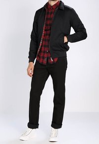 Uomo che indossa una giacca nera sopra una camicia a scacchi rossa e nera, pantaloni neri con risvoltini e sneaker bianche alte, in piedi con una mano in tasca.