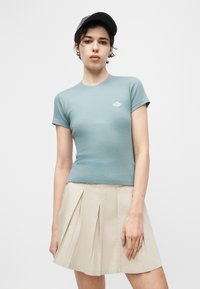 Νεαρό άτομο που φοράει μαύρο καπέλο, ανοιχτό μπλε εφαρμοστό κοντομάνικο πουκάμισο και μπεζ φούστα με πιέτες, στέκεται μπροστά σε απλό λευκό φόντο.