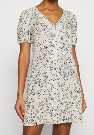 Femme portant une robe blanche à manches courtes avec un motif floral noir et un décolleté en V, tenant ses mains près de ses côtés.
