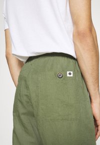 Mand i olivengrønne bukser med en baglomme og knap samt et hvidt mærke, parret med en nedstoppet hvid t-shirt.