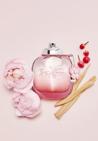 Pink glass perfume bottle with silver cap, featuring an embossed carriage design and a pink flower charm, surrounded by peonies and wood.