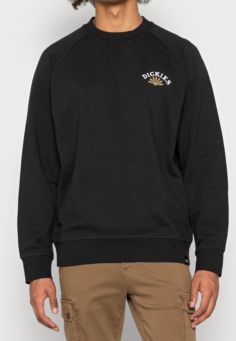 Svart sweatshirt i mjukt tyg med ribbad halsringning, raglanärmar och en broderad logga i vitt och guld på bröstet.