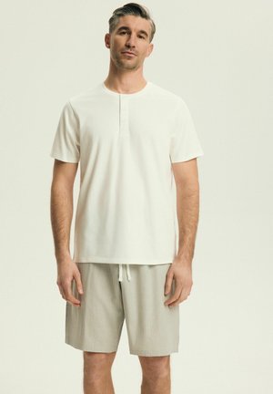 Mężczyzna stojący, ubrany w białą koszulkę henley z krótkim rękawem i jasnoszare spodenki z wiązanym sznurkiem na gładkim beżowym tle.