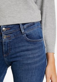 Blå denimjeans med en slät textur, utrustade med en dubbelknäppe stängning, framfickor och kontrasterande sömmar, i kombination med en grå tröja.