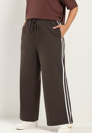 Persoon die donkerbruine wijde broek met witte zijstrepen, witte sneakers en een bruin topje draagt, staand met handen in de zakken.