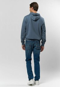 Modrá mikina s kapucňou, kombinovaná s vyblednutými modrými džínami. Tenisky sú biele. Jednoduchý dizajn, bez viditeľných vzorov alebo log. 