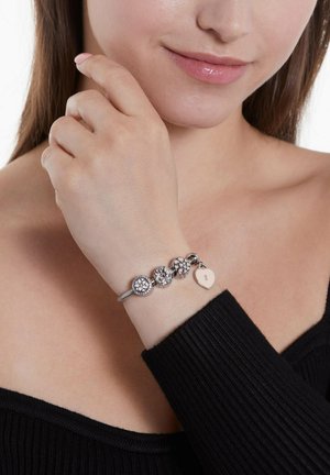 Silbernes Charm-Armband mit drei strukturierten Perlen mit Blumendesigns und einem herzförmigen Charm, getragen an einem Handgelenk mit einem schwarzen Oberteil.