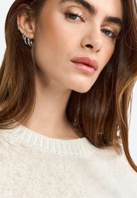 Maglione color crema, con texture e colletto a coste; il modello ha capelli castano medio e indossa numerosi orecchini a cerchio in argento.