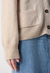 Cardigan en maille beige avec poche avant et boutons porté sur une jupe en denim bleu, montrant le bras gauche et la main posés sur le côté.