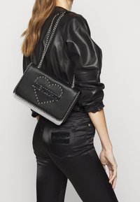 Borsa a tracolla in pelle nera con borchie argentate, design a forma di cuore e tracolla a catena. Abbinata a pantaloni neri lucidi e una giacca nera.