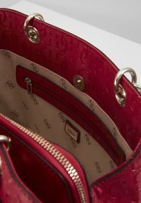 Sac à main rouge avec extérieur texturé, ferrures dorées et une poche zippée. L'intérieur présente une doublure beige avec un motif de logo discret.