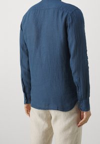 Hombre vestido con una camisa de lino azul oscuro de manga larga y pantalones de lino beige claro, visto desde atrás contra un fondo blanco liso.