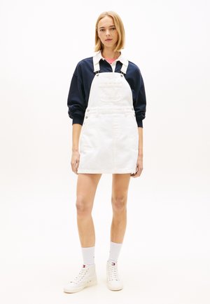 Jeune femme debout portant une robe salopette en denim blanc sur un sweat-shirt bleu marine, des chaussettes blanches et des baskets montantes blanches sur un fond blanc.