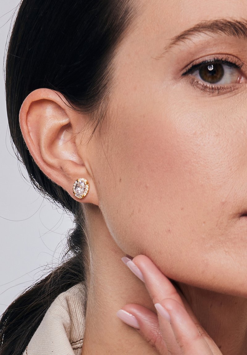 Złote, studowe kolczyki z dużym owalnym przezroczystym kamieniem otoczonym mniejszymi przezroczystymi kamieniami, osadzonymi w teksturowanej metalowej bazie.