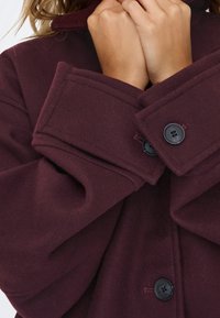 Mains agrippant le col d'un manteau en laine violet foncé avec de grands boutons noirs et de larges poignets.