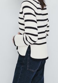 Sweter w paski w czarno-białym wzorze, z prążkowanymi mankietami, bocznymi rozcięciami i luźnym krojem. W połączeniu z ciemnoniebieskimi dżinsami.