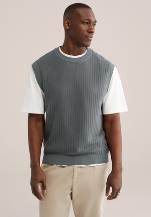 Muž v šedej pletenej bezrukávovej veste cez bielu košeľu s krátkym rukávom a béžových nohaviciach, pozerajúci sa do strany na jednoduchom pozadí.