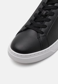 Adidași negri din piele cu vârful rotunjit, partea superioară texturată, talpă din cauciuc alb și șireturi negre. Detalii de cusătură vizibile pe părțile laterale.