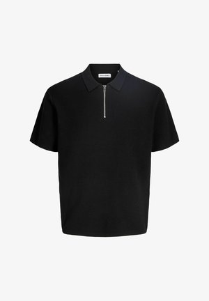 Uśmiechnięty chłopiec stoi, ubrany w czarną koszulkę polo z krótkim rękawem, szare dżinsy i białe trampki na jednolitym jasnym tle.